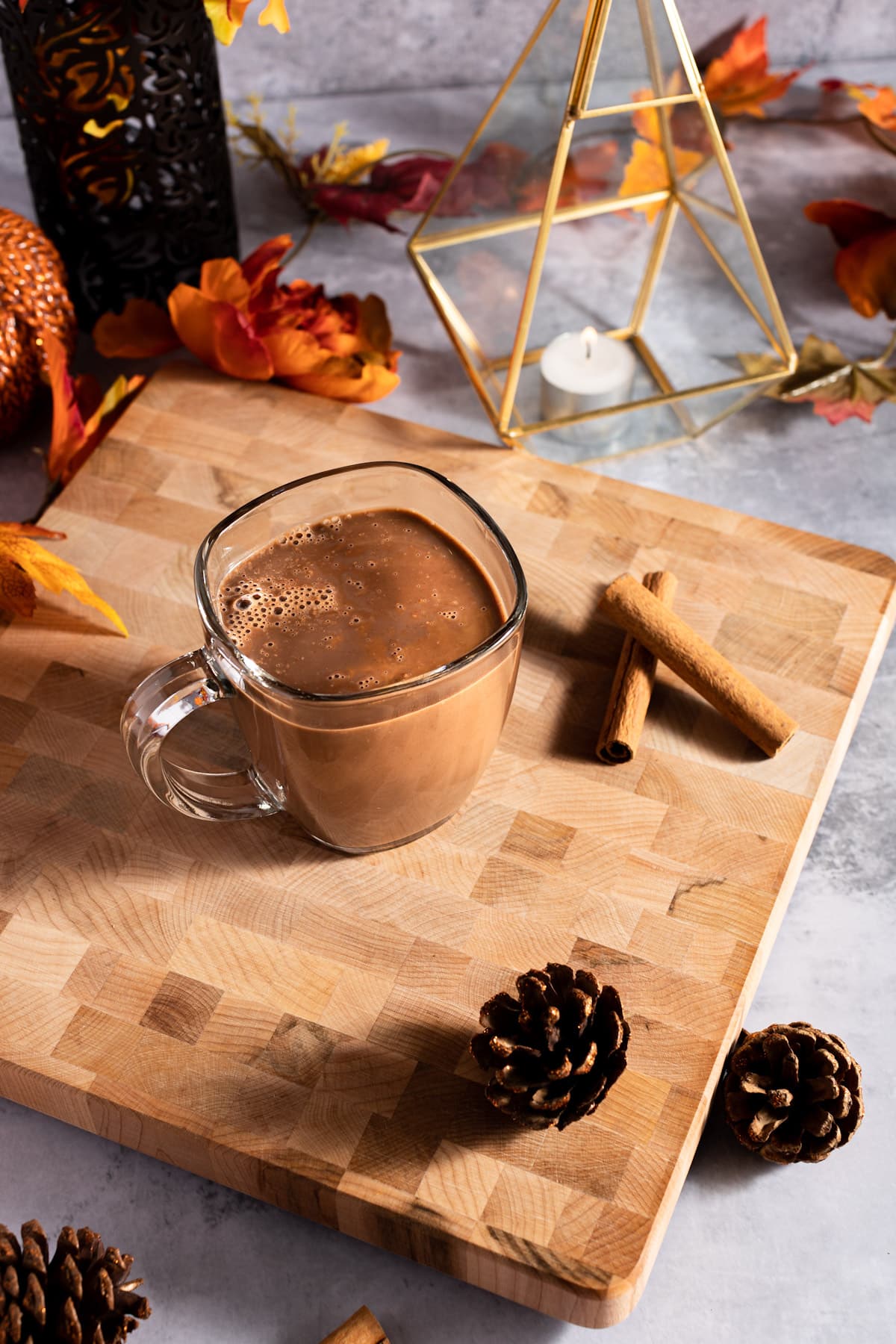 Overhead photo of oat hot chocolate, next to pinecones, cinnamon sticks, a small candle in a golden votive and leaves.