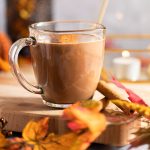Oat milk hot chocolate on a wooden serving board, next to cinnamon sticks, with fall leaves in the foreground.