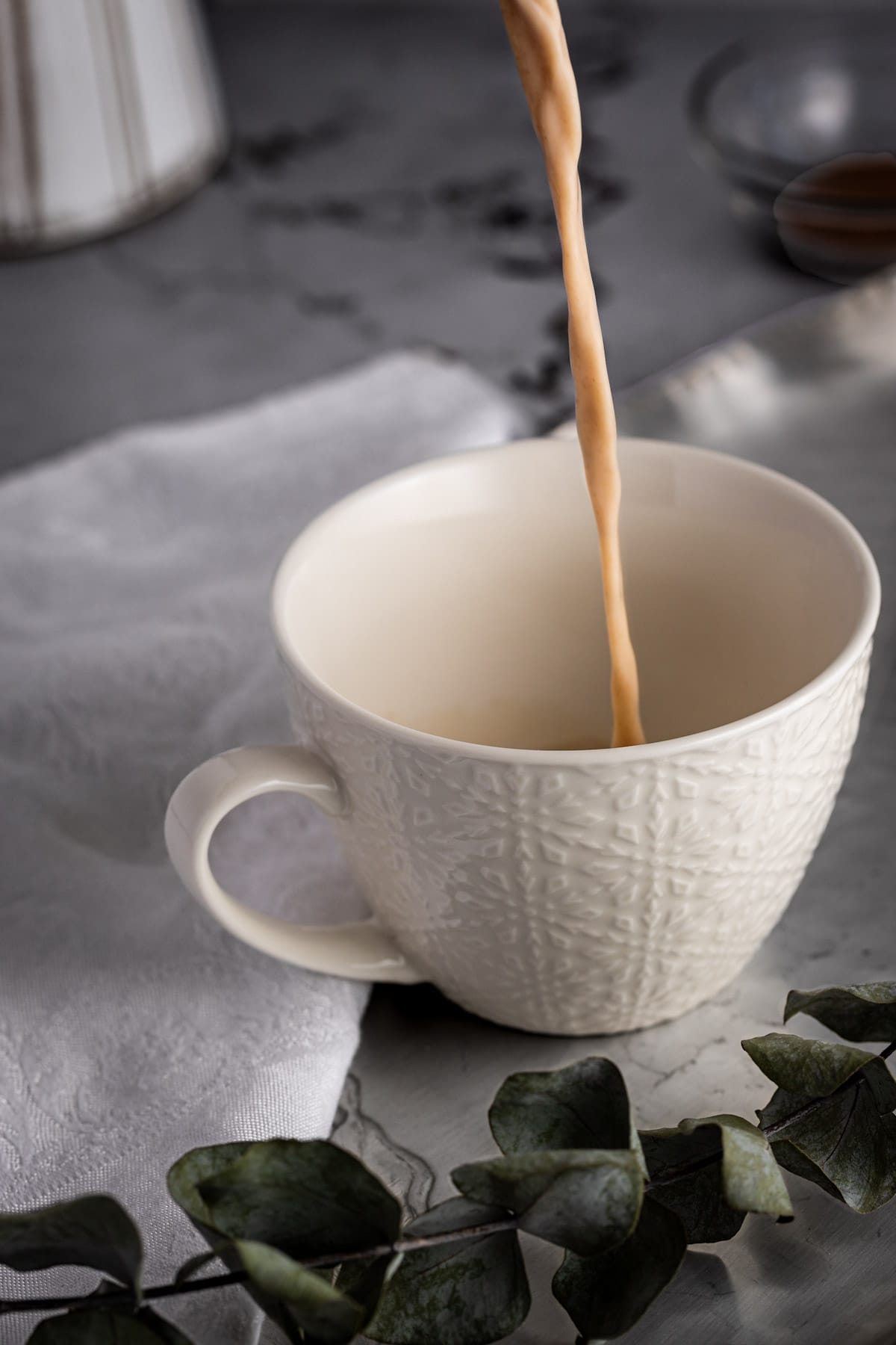 Oat milk latte being poured into a white coffee mug, next to a white linen napkin.