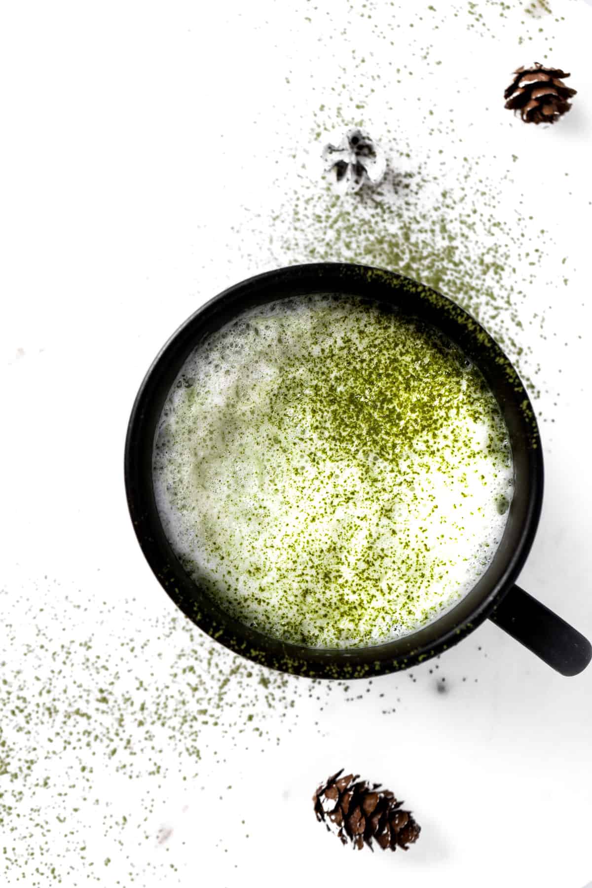 A cup of oat milk matcha latte in a black mug, sprinkled with matcha powder on a white table.