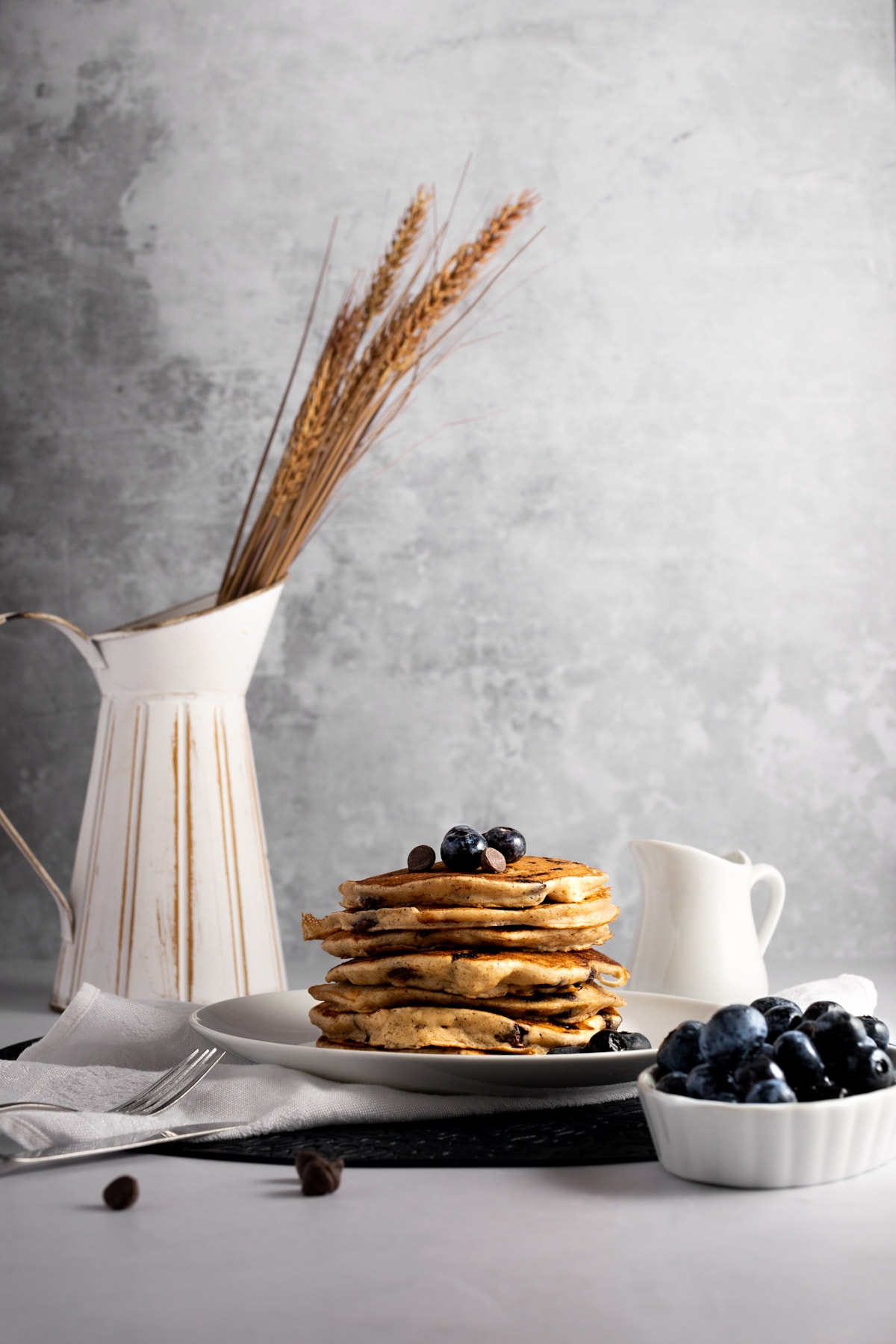 A stack of pancakes make with oat milk on a white plate, next to a white dish filled with fresh blueberries, a container with maple syrup and a rustic just with wheat.