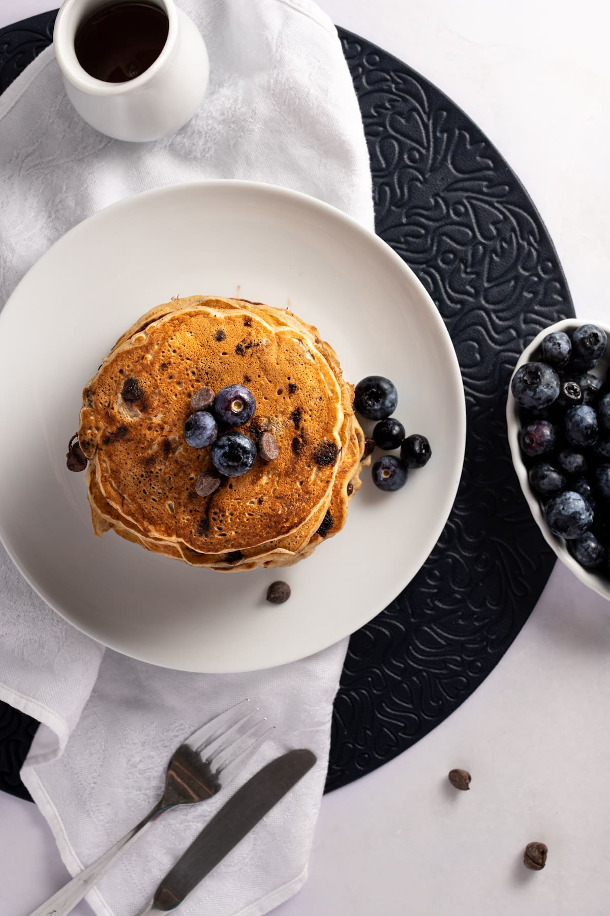 Overhead view of a stack of pancakes topped with blueberries, sitting on a white plate with a round blue placemat, white napkin and silver fork and knife.