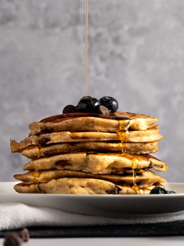 Maple syrup being poured over a stack of oat milk pancaked topped with blueberries and chocolate chips, sitting on a white plate, with a grey background.