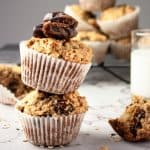 Up close side shot of two banana date oat muffins balanced on top of each other with milk and more muffins stacked in the background.