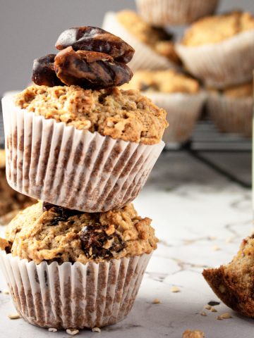 Up close side shot of two banana date oat muffins balanced on top of each other with milk and more muffins stacked in the background.