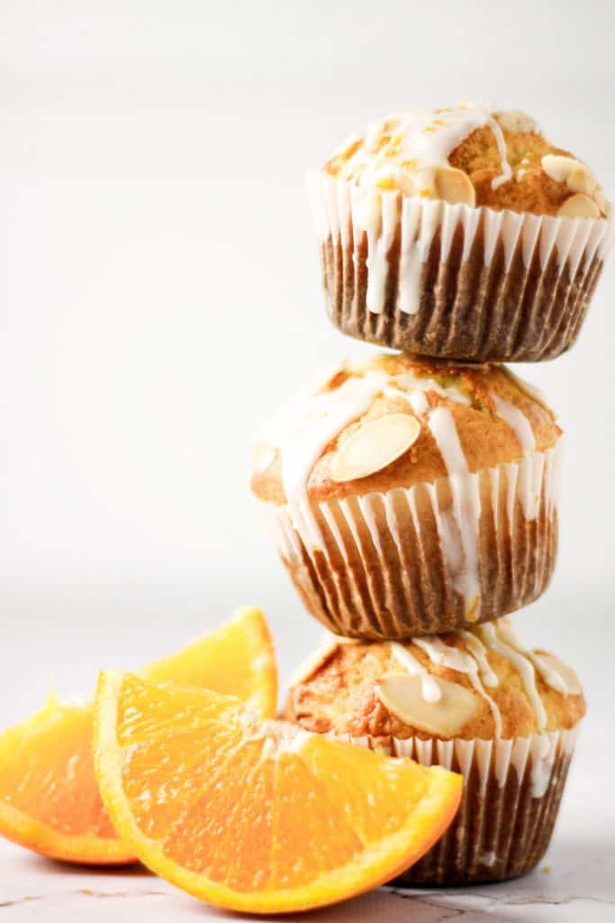 Three orange almond muffins with orange glaze stacked on top of each other next to two orange slices
