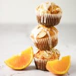 Three orange almond muffins with orange glaze stacked on top of each other next to two orange slices