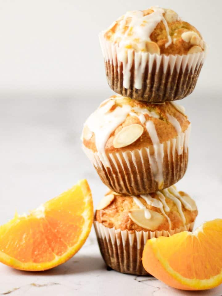 Three orange almond muffins with orange glaze stacked on top of each other next to two orange slices