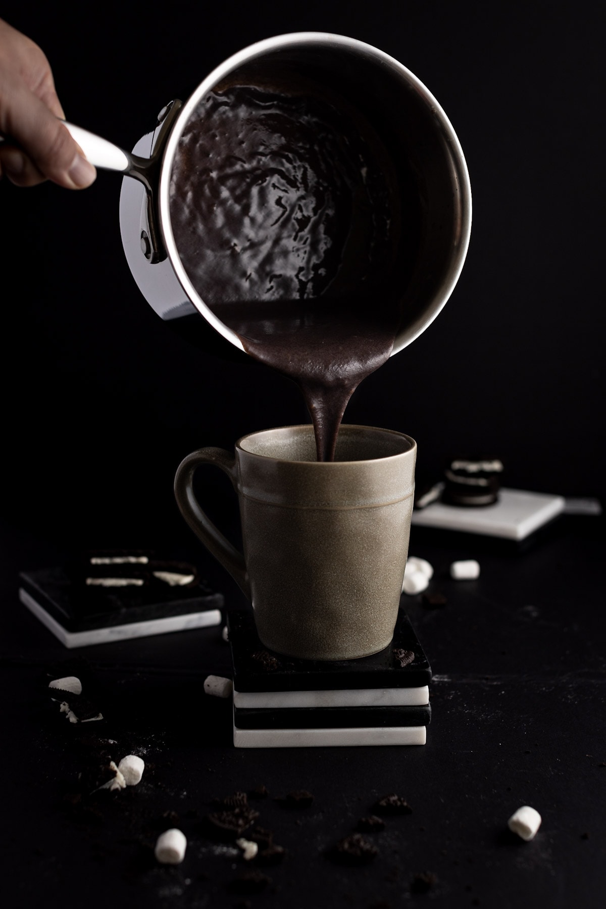 Oreo hot chocolate being poured from a metal saucepan into an olive green mug, on a stack of black and white coasters, with a black background.