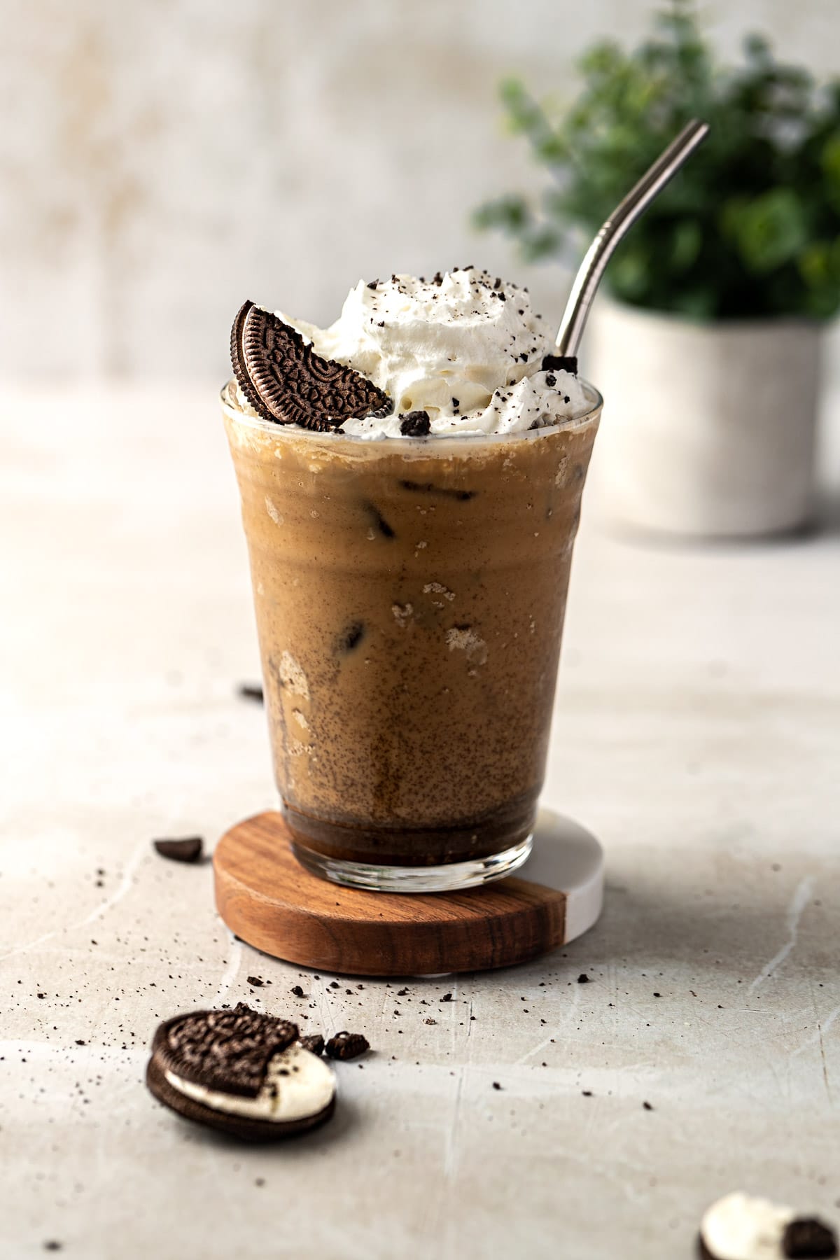 An oreo iced coffee served with a metal straw, topped with whipped cream and oreo chunks, sitting on a warm marble table.