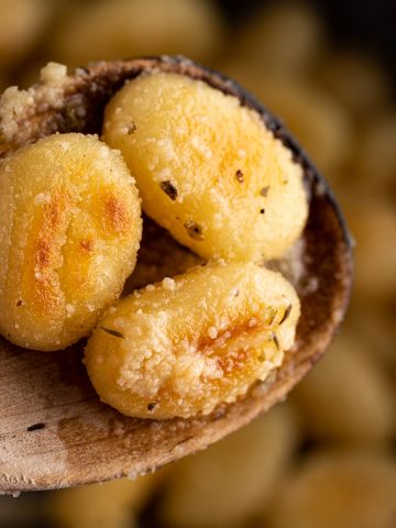 Close up view of 3 pieces of crispy pan fried gnocchi covered in garlic parmesan sauce on a wooden spoon.