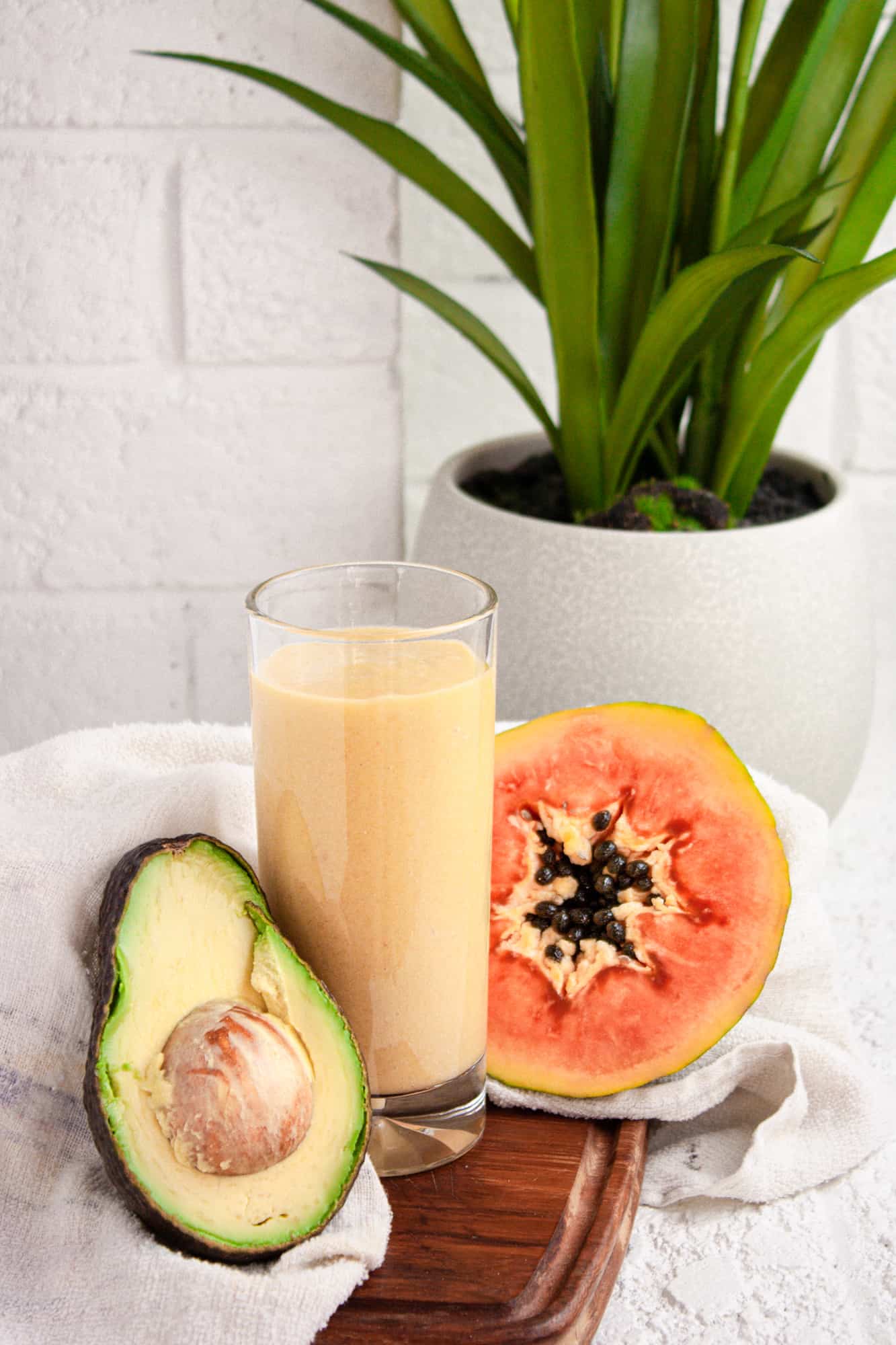 Tall glass of papaya avocado smoothie on a wooden board beside half an avocado and papaya, in front of a white brick wall