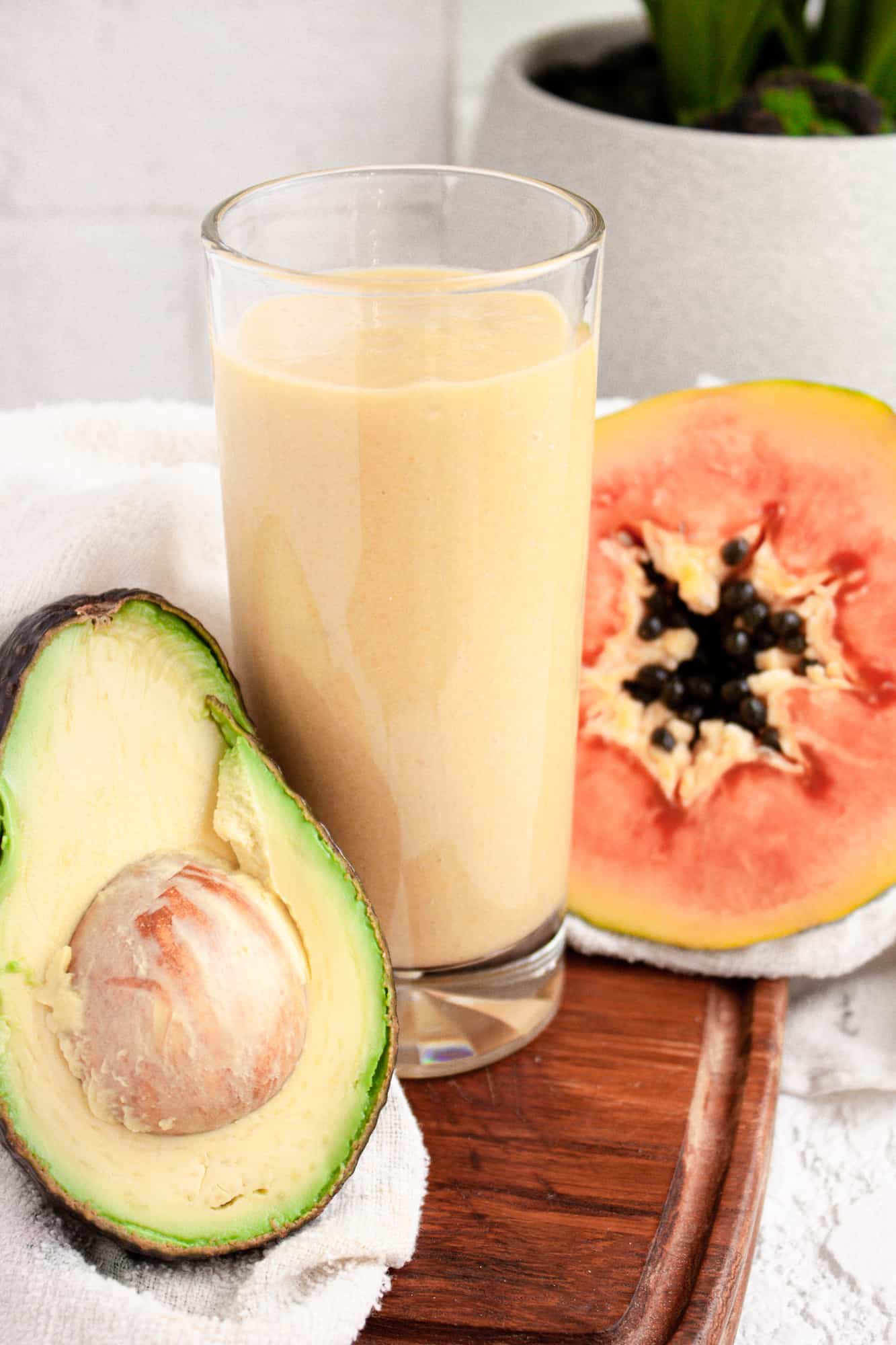 Up close view of a glass of papaya avocado smoothie on a wooden board beside half an avocado and papaya