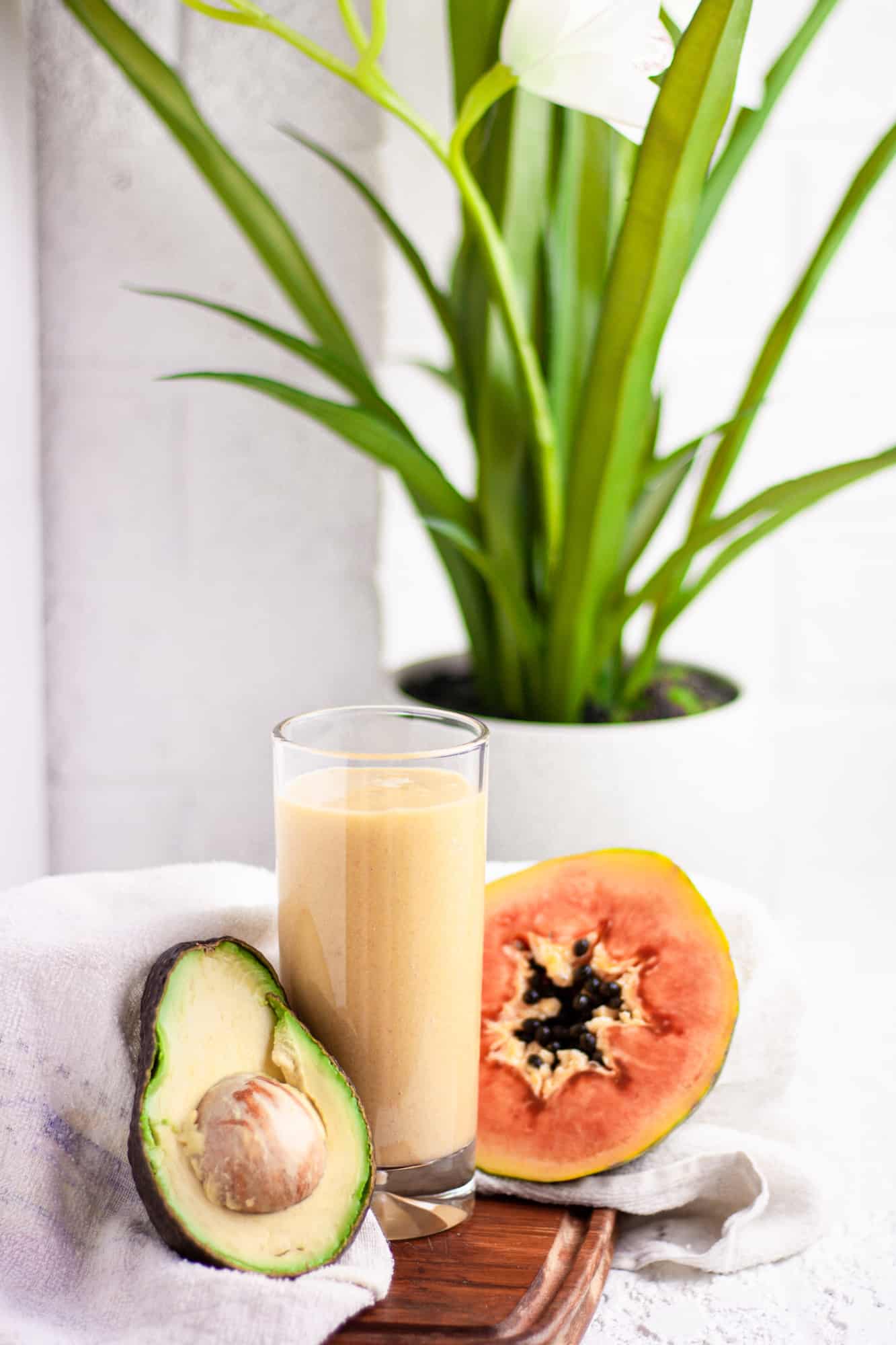 Tall glass of papaya avocado smoothie on a wooden board beside half an avocado and papaya, in front of a white brick wall