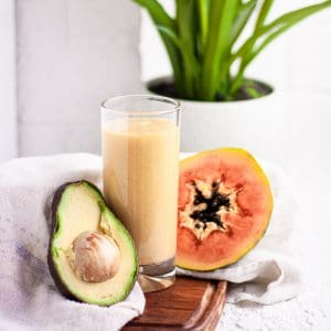 Tall glass of papaya avocado smoothie on a wooden board beside half an avocado and papaya, in front of a white brick wall