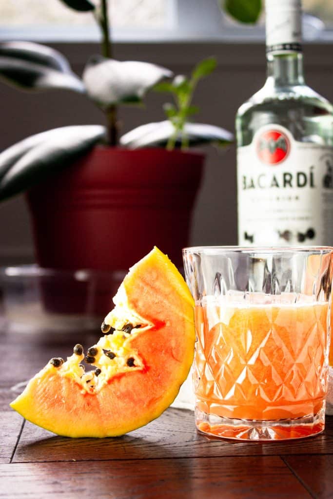 Glass of papaya rum punch beside a papaya slice with a rum bottle and green plant in the background