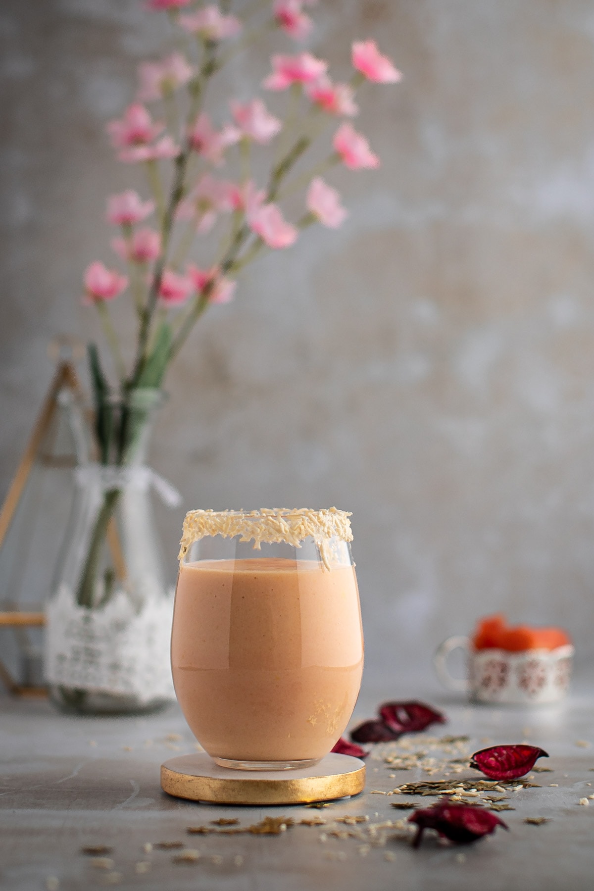 A glass of light pink papaya smoothie on a gold coaster, with pink flowers in the background.