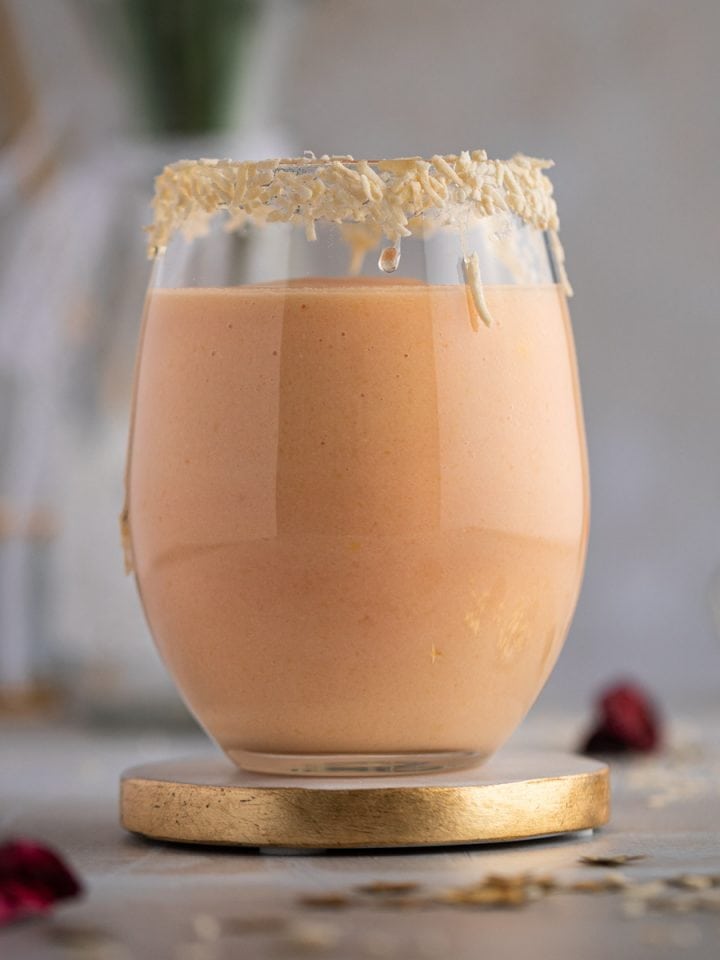 A light pink colored papaya smoothie in a large round glass, with the rim of the glass lined with shredded coconut.