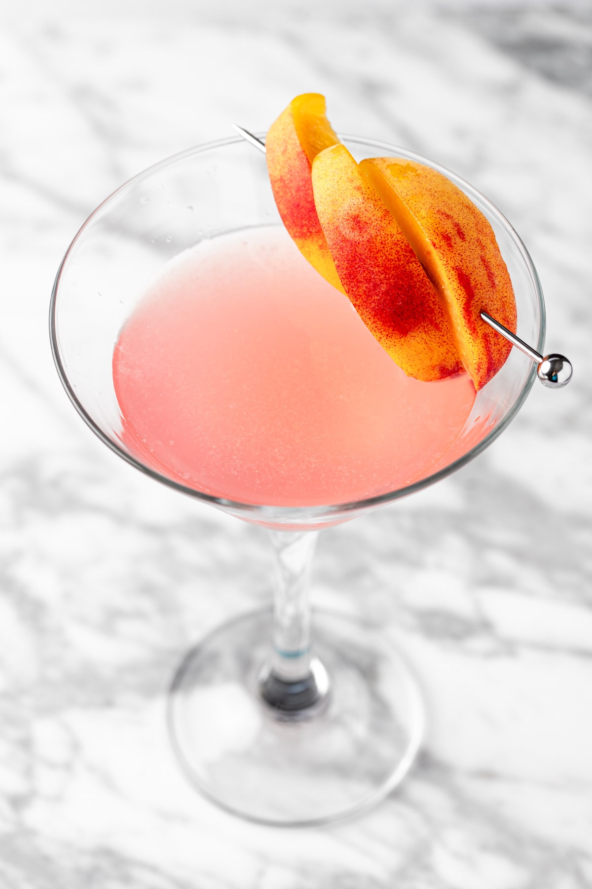 A pink peach cosmopolitan garnished with peaches, on a white marble table.
