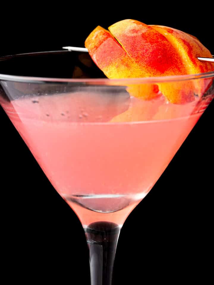 A peach cosmo garnished with peach slices, on a black background.