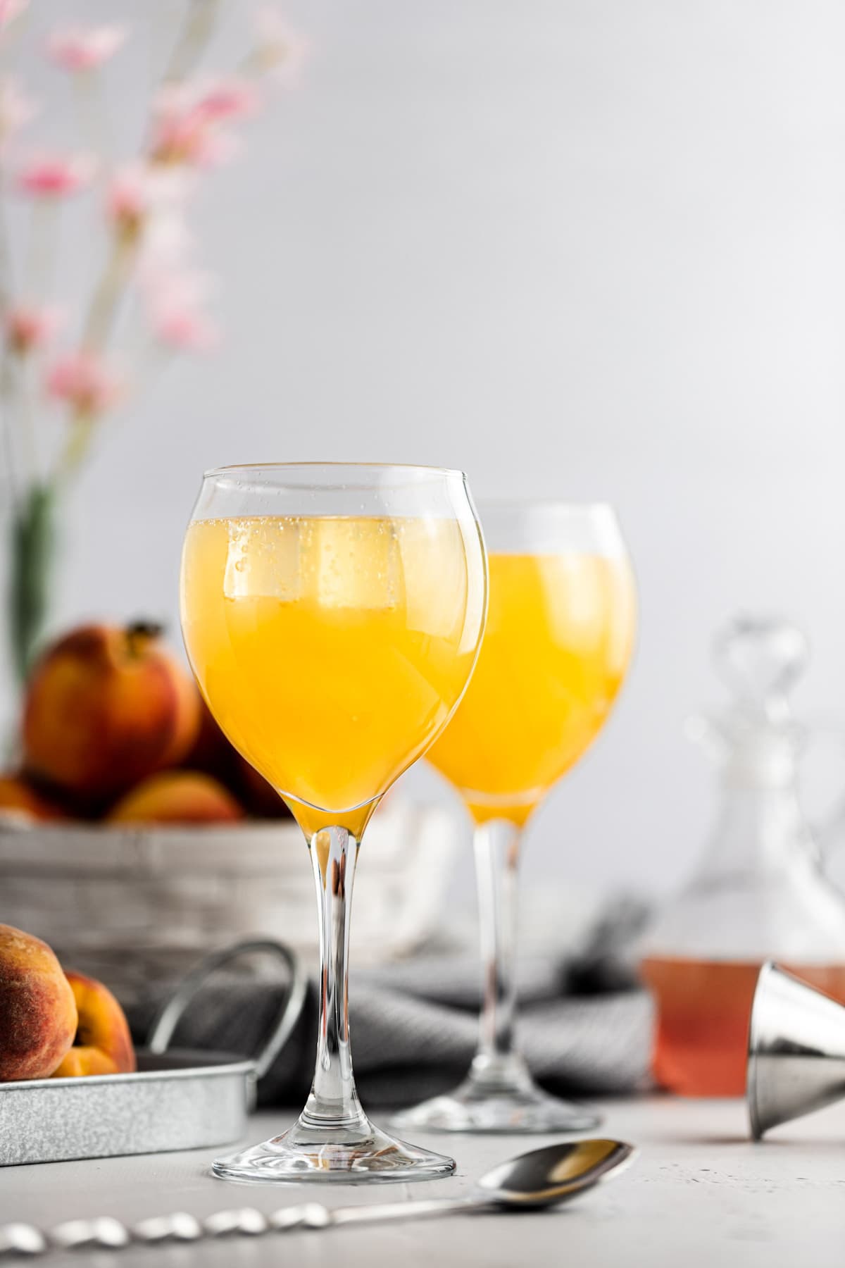 Two peach gin cocktails, next to a basket of fresh peaches, with a grey background.