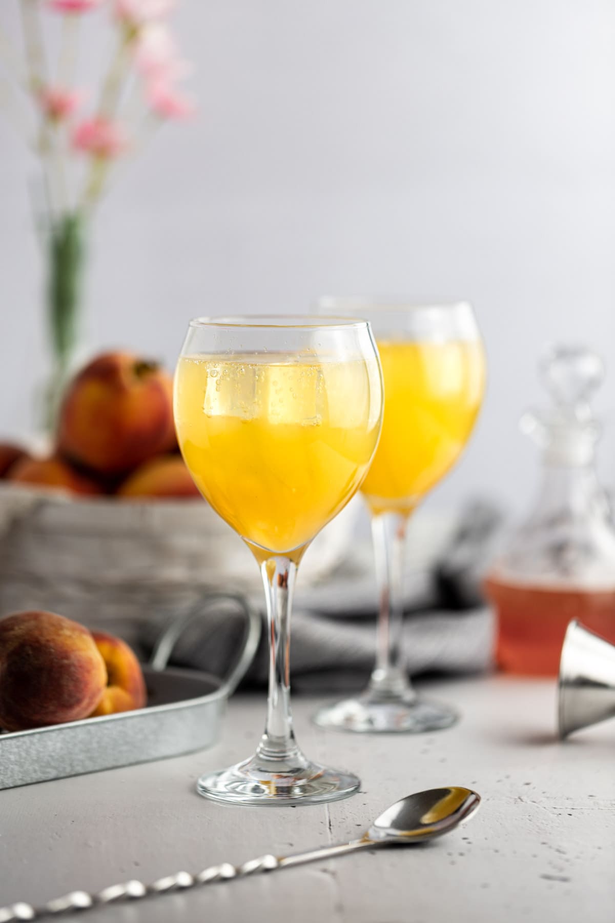 Two peach cocktails on a grey table, next to fresh peaches and a jar of peach syrup.