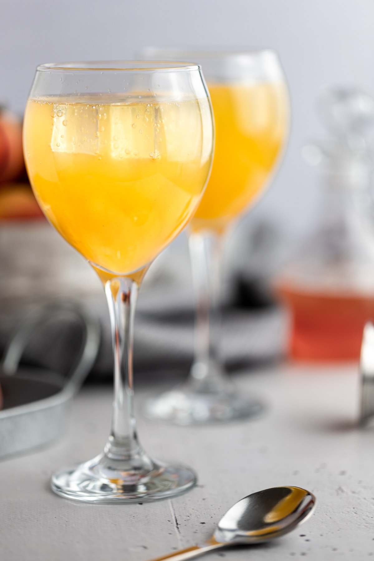 Up close view of an orangey-yellow gin peach cocktail, next to a spoon and fresh peaches with a grey background.