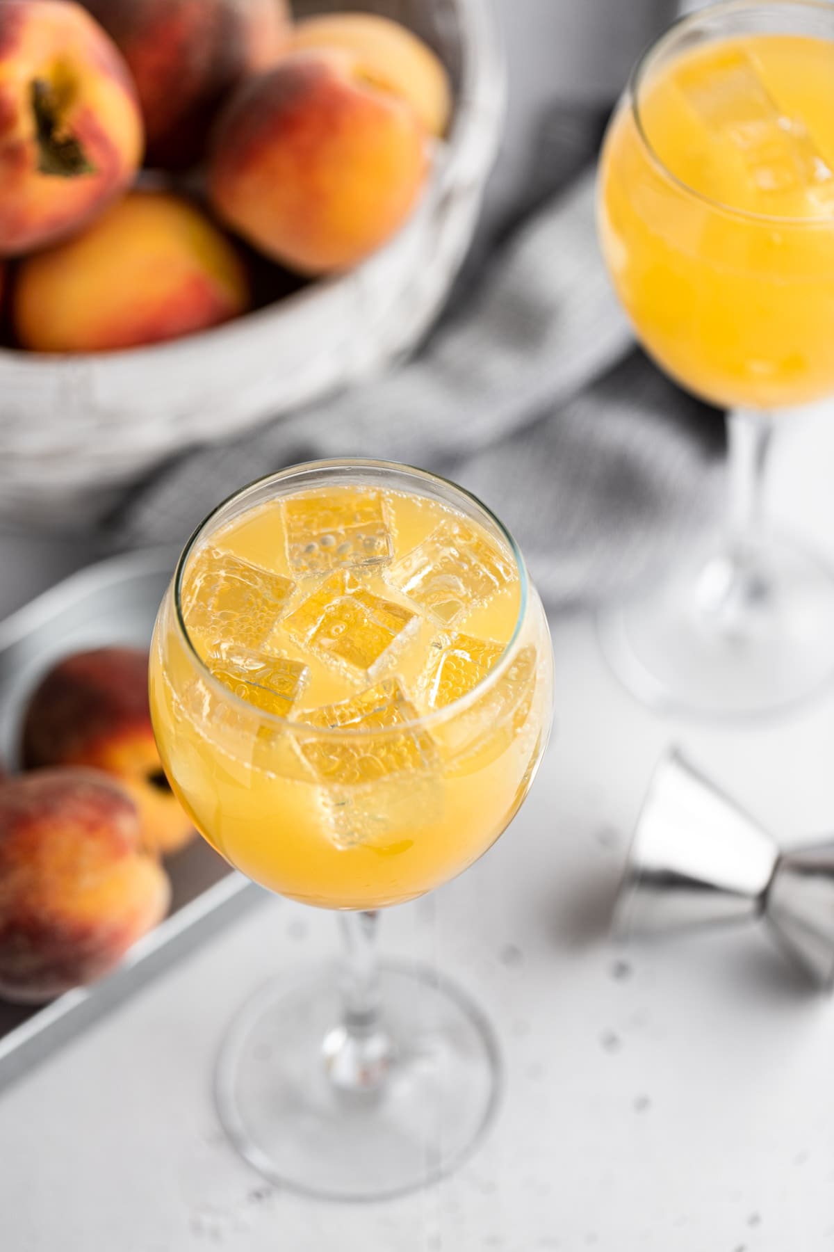 Overhead view of a peach gin cocktail with ice cubes, on a light grey table, next to a metal jigger and metal tin filled with fresh peaches.