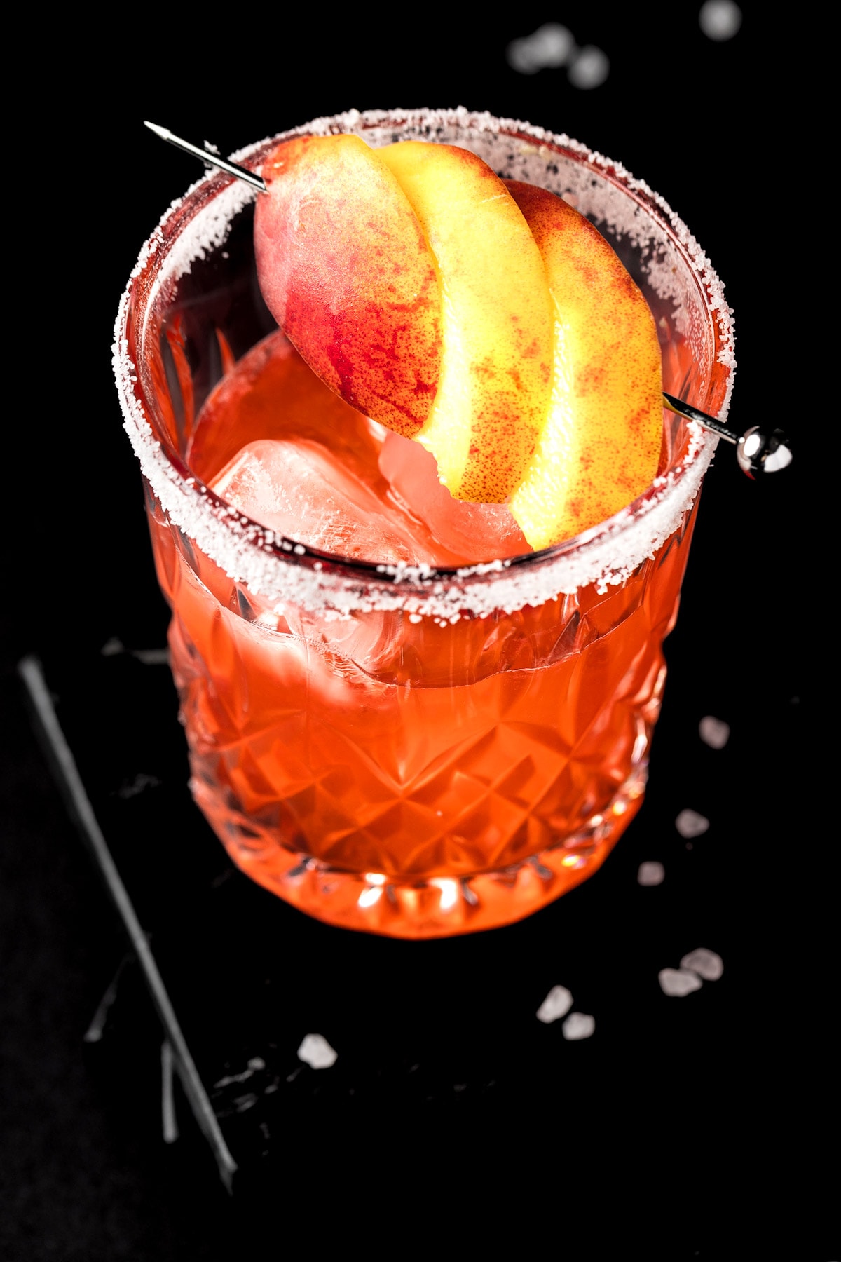 A peach margarita on the rocks garnished with peach slices on a dark table.