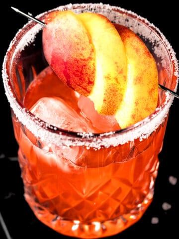 A peach margarita on the rocks garnished with peach slices on a dark table.