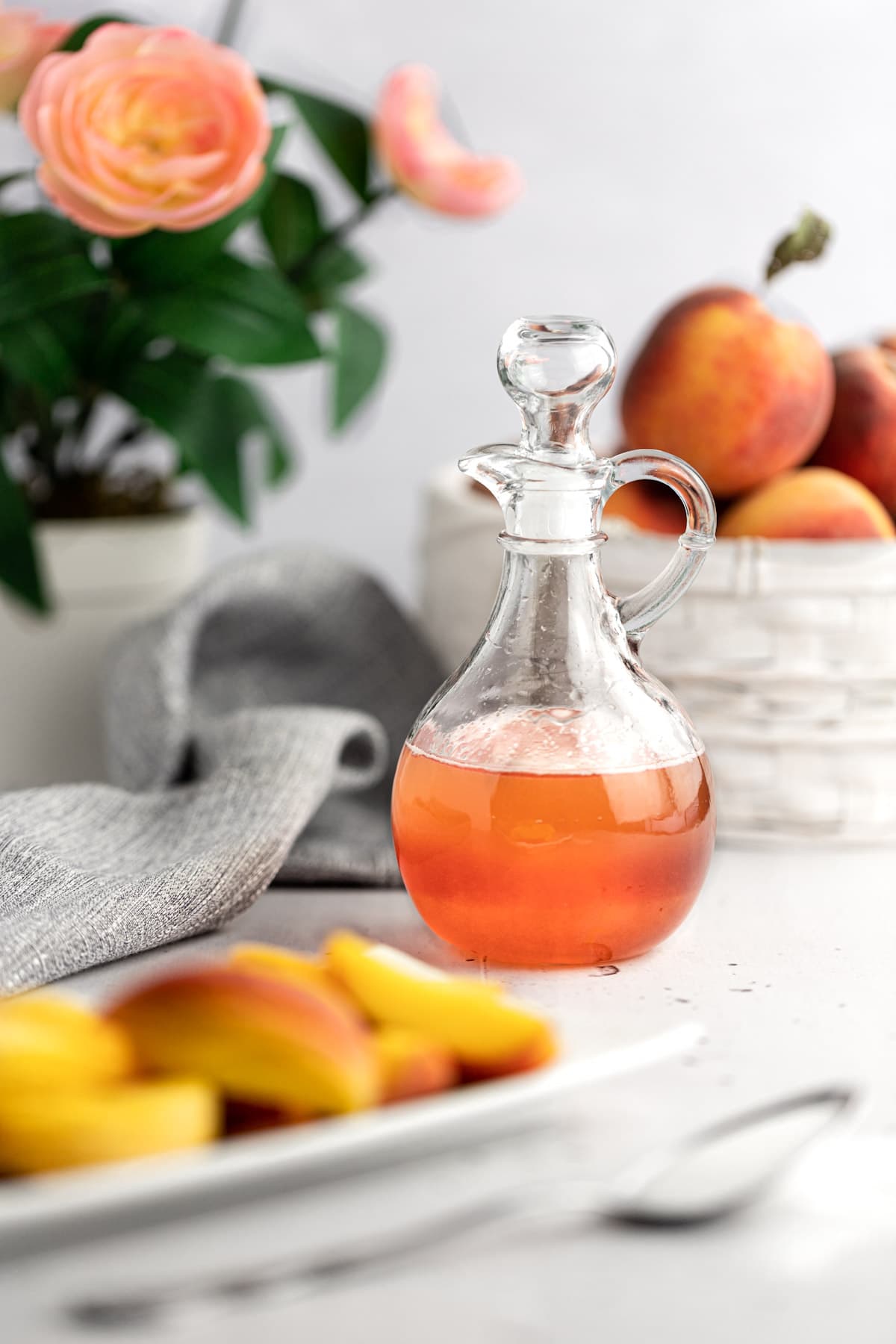 Peach syrup jar on a white table next to peach slices, a basket of peaches and pink flowers.