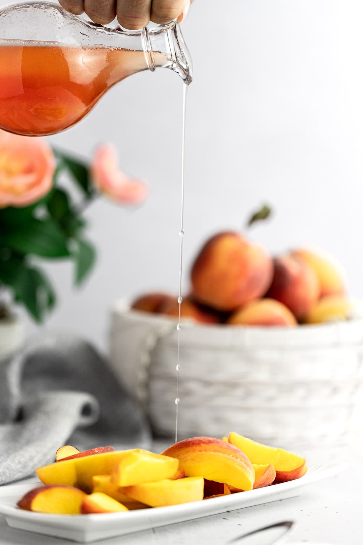 Syrup being poured over a plate of fresh peach slices, a grey napkin and basket of peaches in the background.