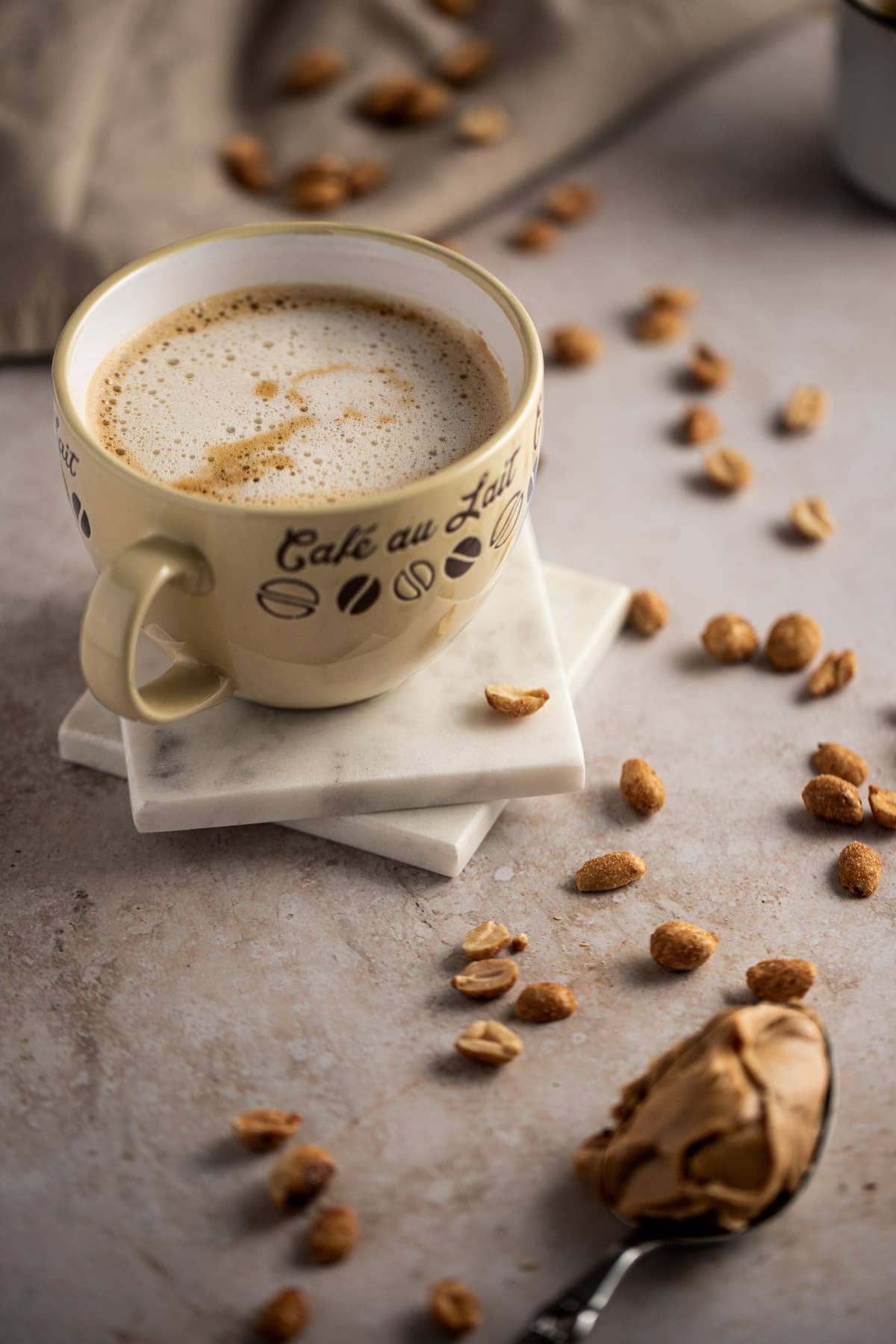 Peanut butter latte in a yellow cappuccino mug, on a couple white marble coasters, next to scattered peanuts.