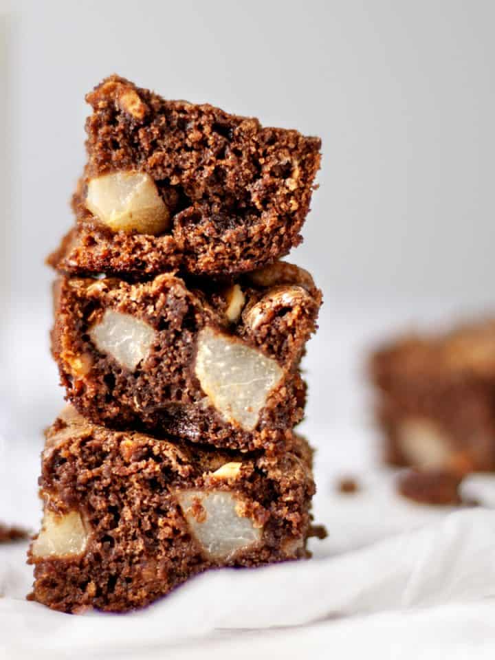 Three pear and hazelnut brownies stacked on top of each other, sitting on parchment paper