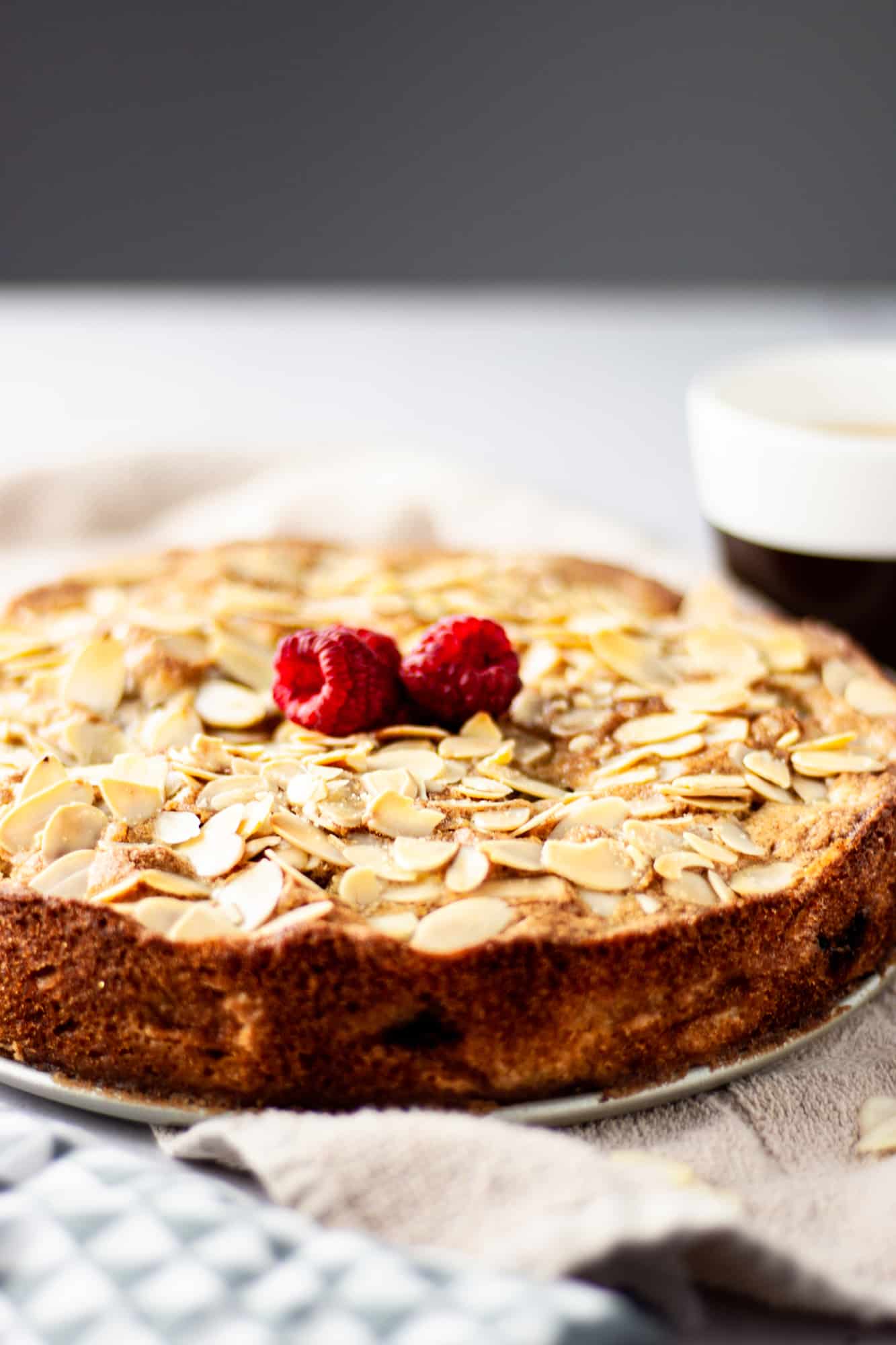 A fresh out of the oven pear and raspberry cake with sliced almonds and raspberries on top next to a cup of coffee.