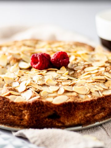 A fresh out of the oven pear and raspberry cake with sliced almonds and raspberries on top next to a cup of coffee.