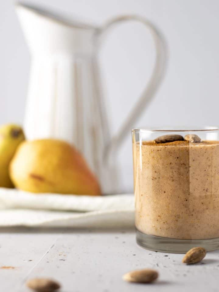 Side view of a glass of pear banana smoothie with three almonds in front and pears and a white jug in the background