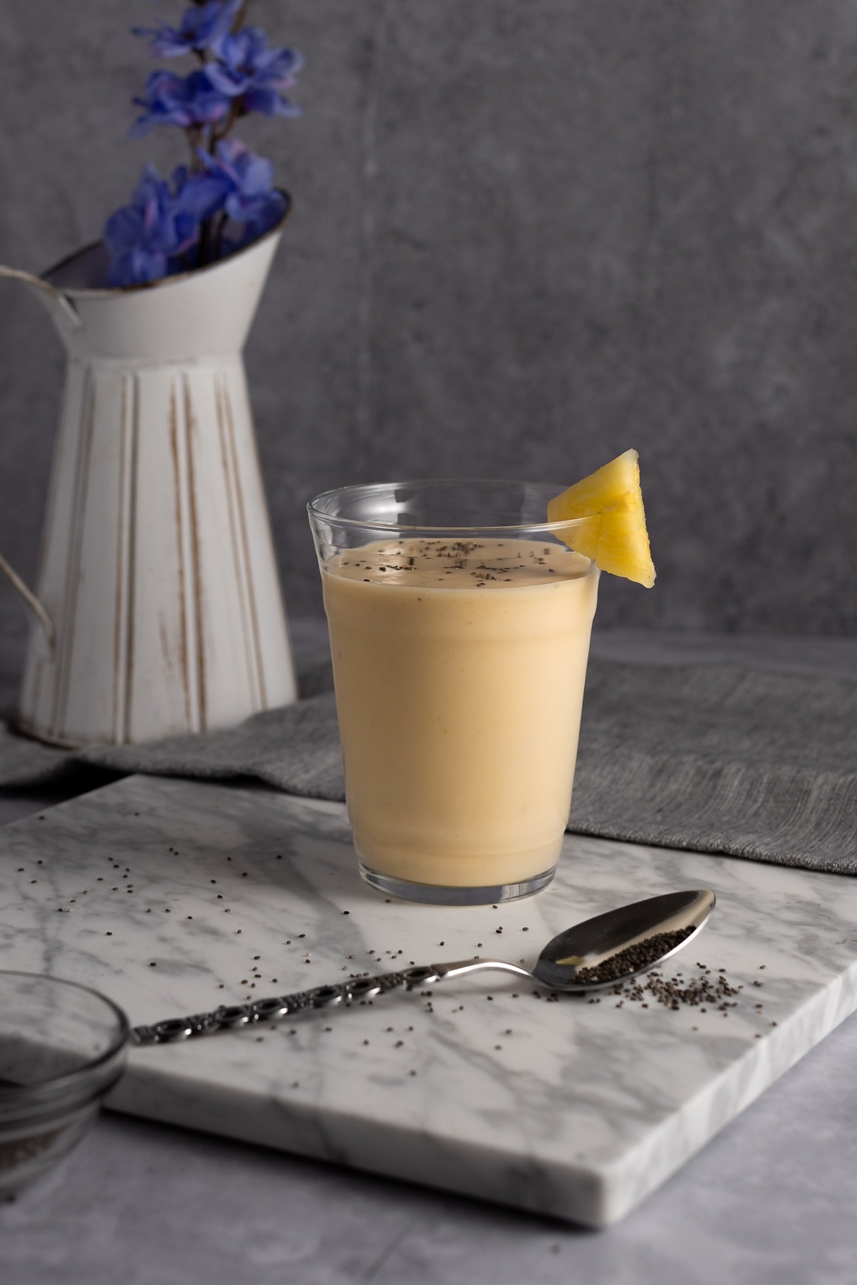 A yellow pineapple banana smoothie on a grey marble board, next to a grey napkin, with a white pot filled with blue flowers in the background.