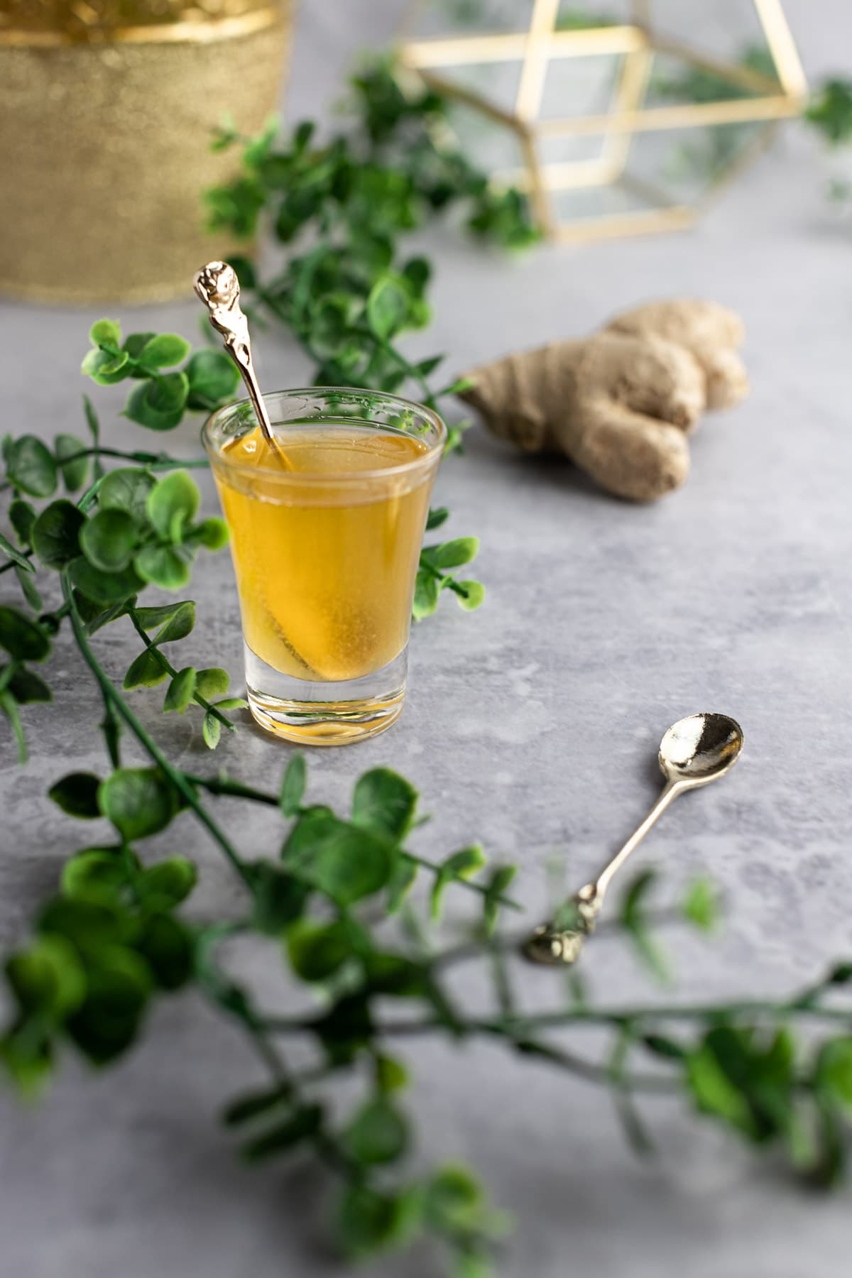 A small glass of simple syrup, on a grey table, next to a golden spoon, with green leaves across the table.