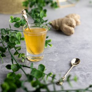 A small glass of golden pineapple ginger syrup, next to a small spoon, on a grey table, with green vines spread across the table.