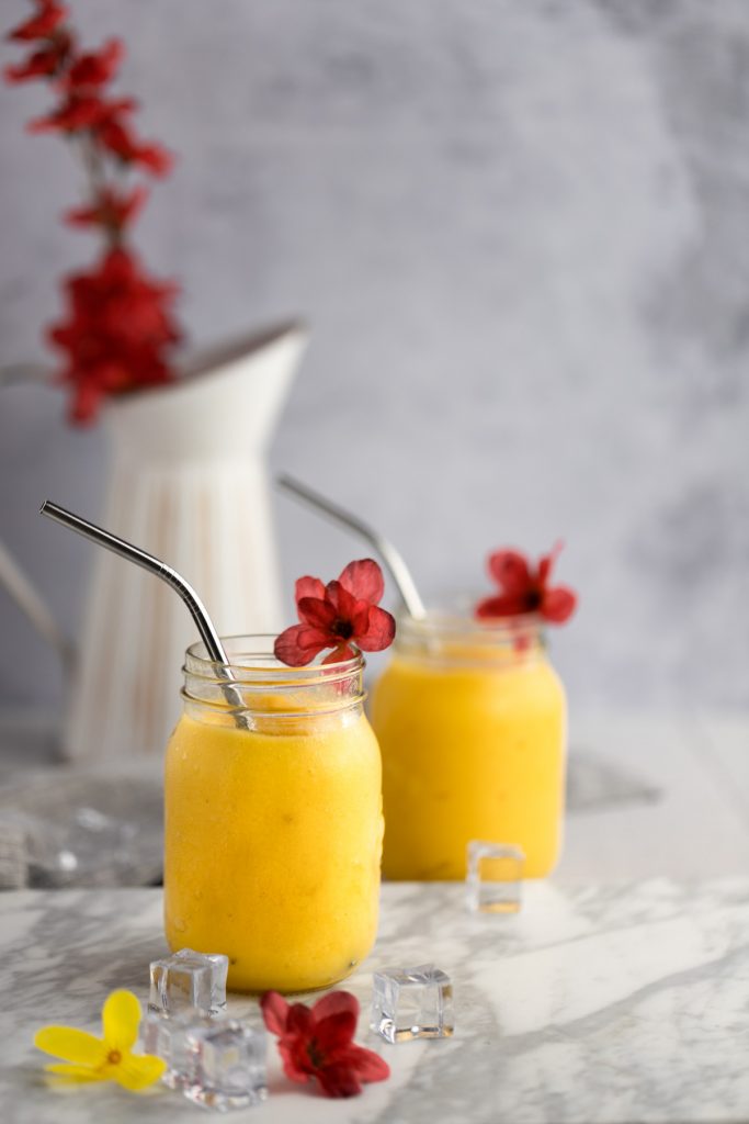 A couple mango pineapple smoothies with metal straws, red flowers on edge of glass, with white vase filled with more red flowers in the background.