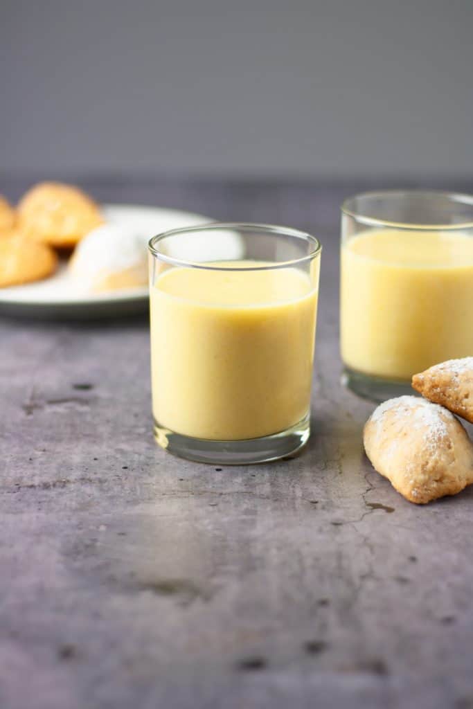 A glass of yellow pineapple mango smoothie on a table with a couple of pastries in front of the glass with pastries in the background.