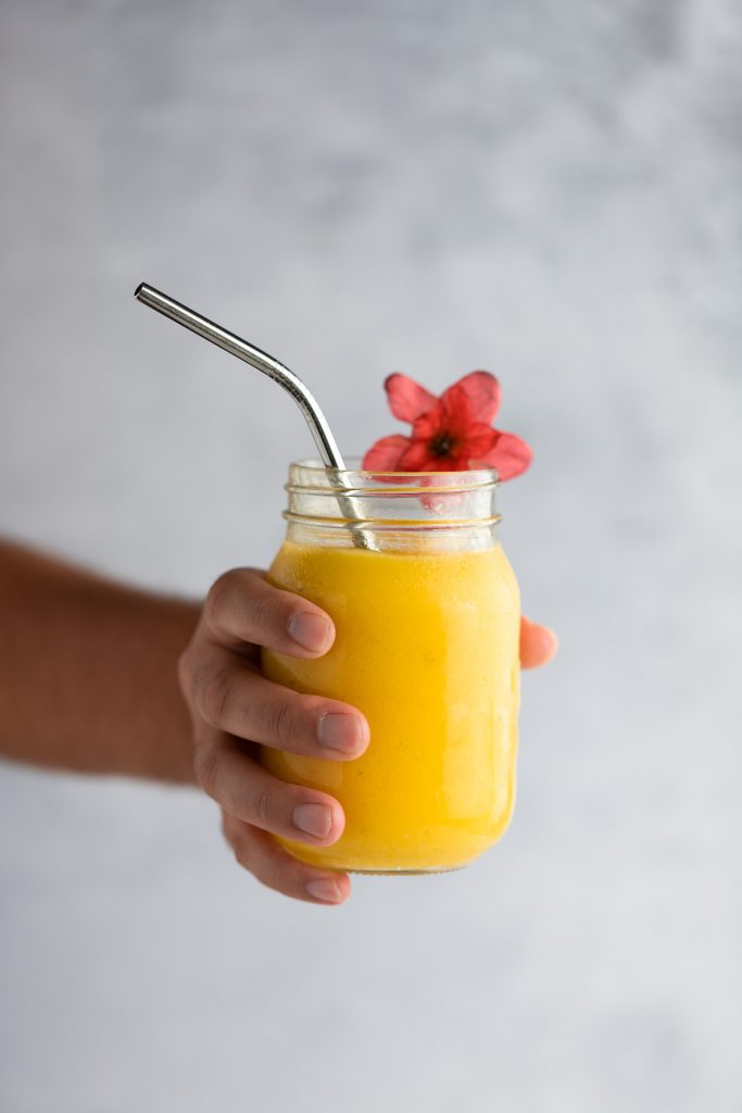 A hand holding a pineapple mango smoothie with a metal straw and red flower on the edge, in front of a grey background.