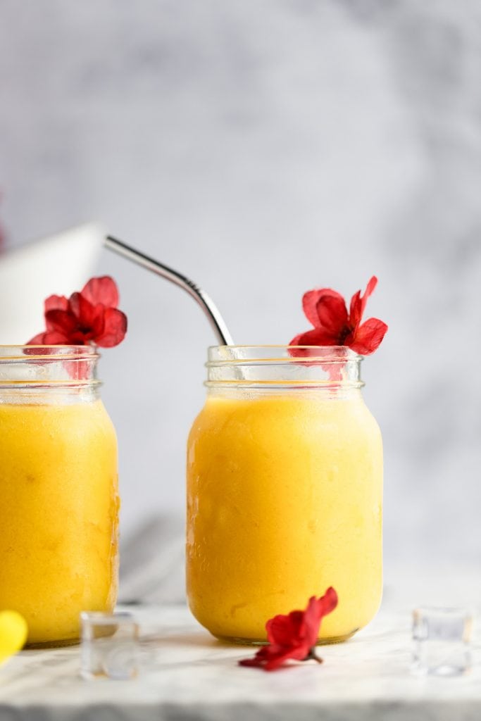 Two pineapple mango smoothies, one partially cut out of the frame, with red flowers and ice cubes scattered at the bottom of the jars.