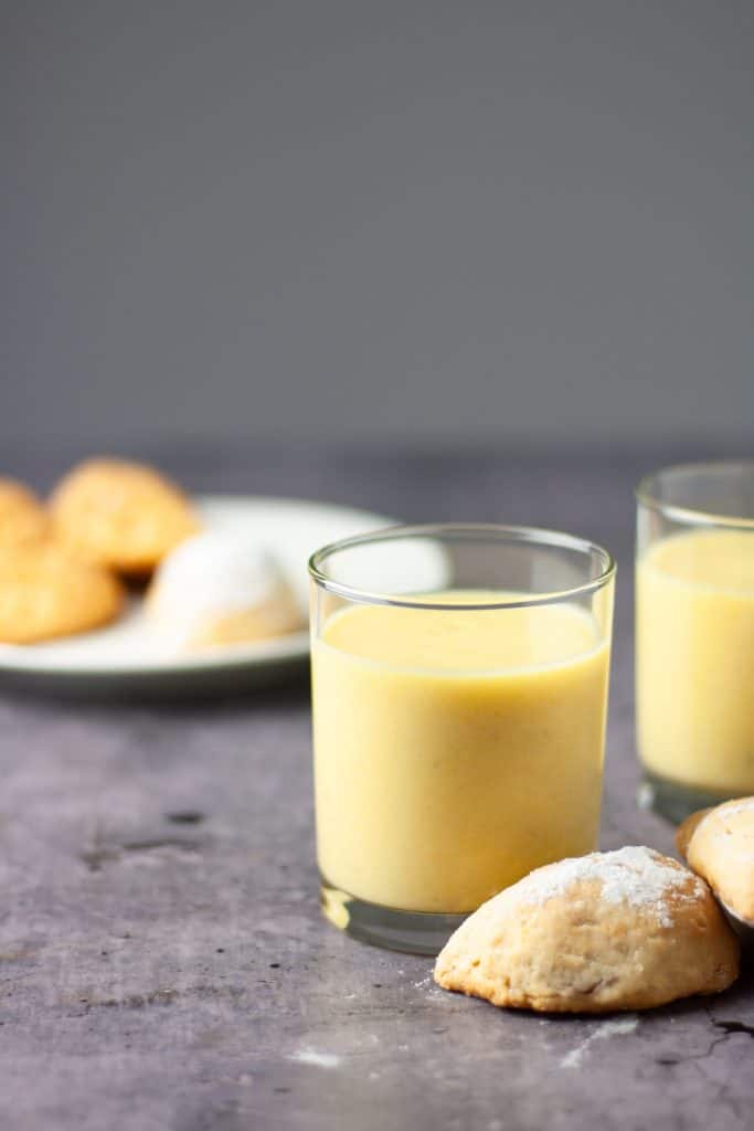 A glass of yellow pineapple mango smoothie on a table with a couple of pastries in front of the glass and a plate of pastries in the background