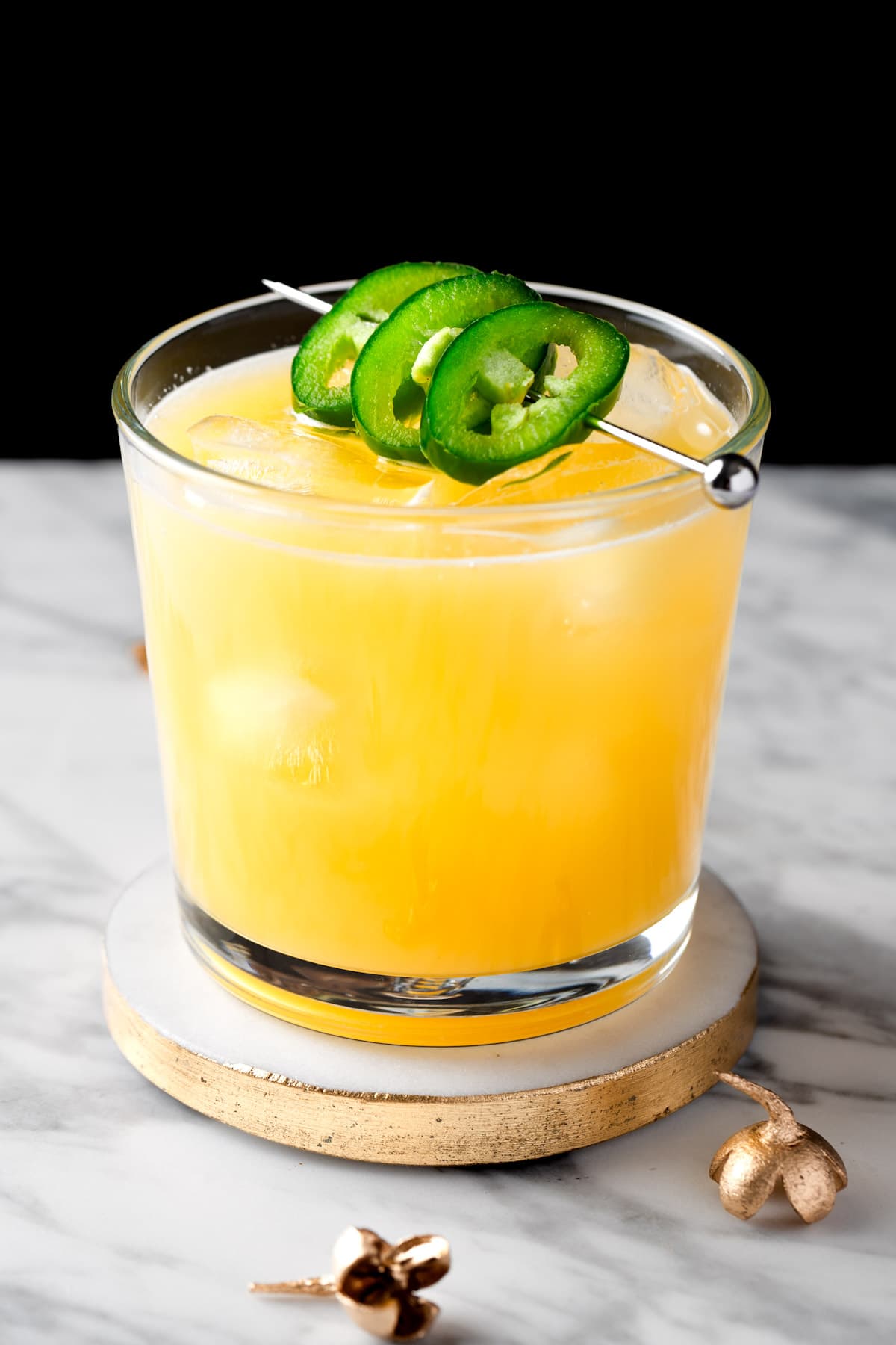 A pineapple mezcal cocktail garnished with jalapeno slices, on a white and black background.
