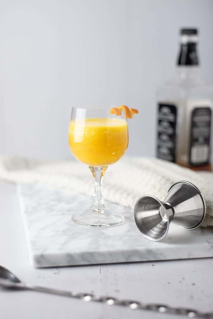 A pineapple old fashioned on a marble cutting board with a spoon in front and jigger and whiskey bottle in the background