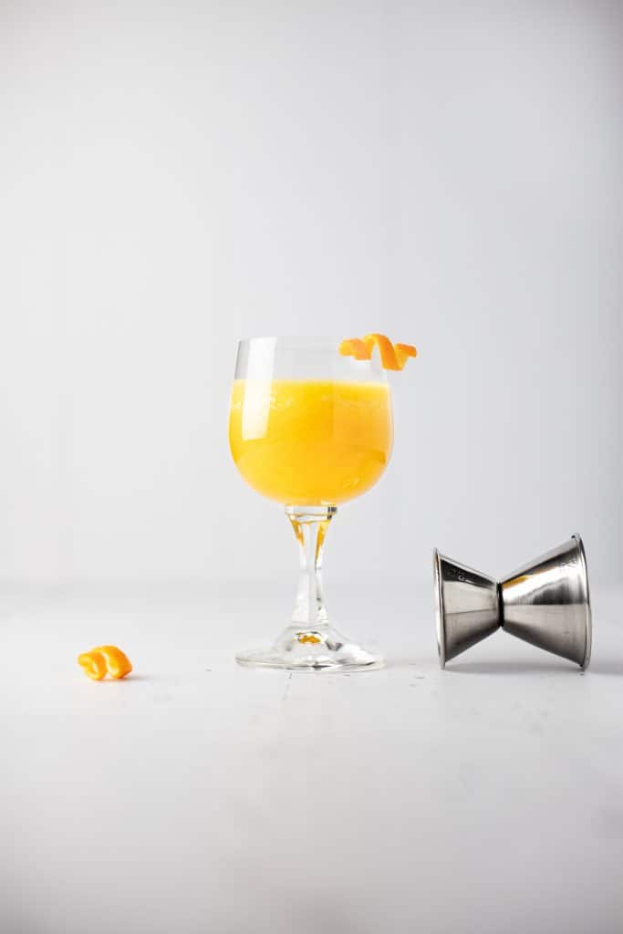 A pineapple old fashioned cocktail with an orange peel garnish next to a toppled jigger and extra orange garnish