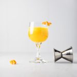 A pineapple old fashioned cocktail with an orange peel garnish next to a toppled jigger and extra orange garnish