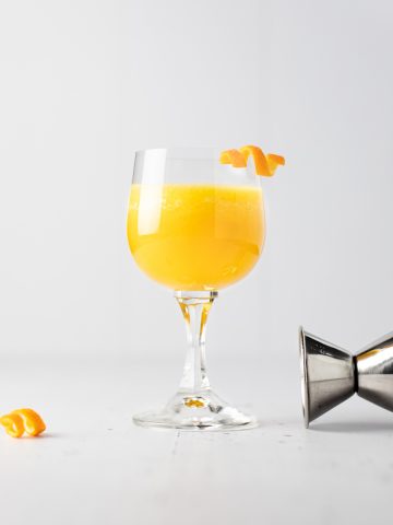 A pineapple old fashioned cocktail with an orange peel garnish next to a toppled jigger and extra orange garnish